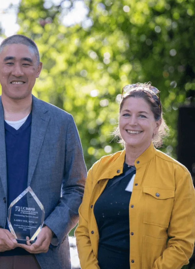 Three people stand outdoors smiling; the person in the center holds a clear award plaque. Trees and sunlight are visible in the background.