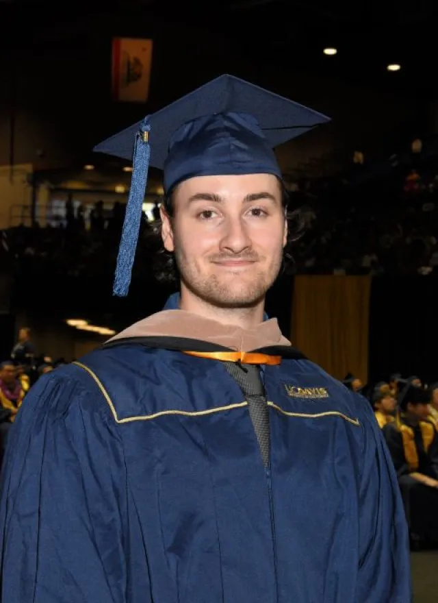 A person in a blue graduation cap and gown stands indoors at a graduation ceremony, with other graduates and an audience visible in the background.