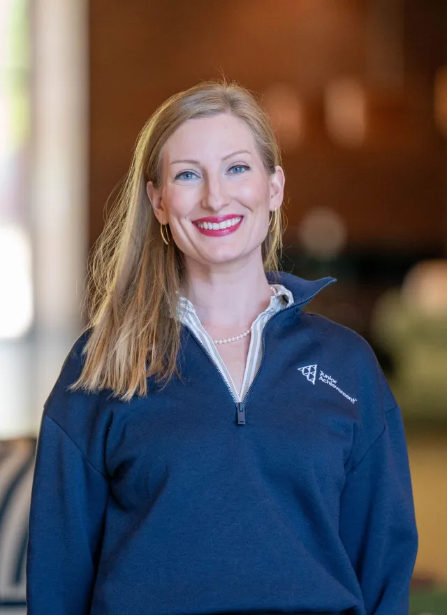 Jaimie Asakawa smiling, wearing a navy pullover and pearl necklace, standing indoors with blurred background.