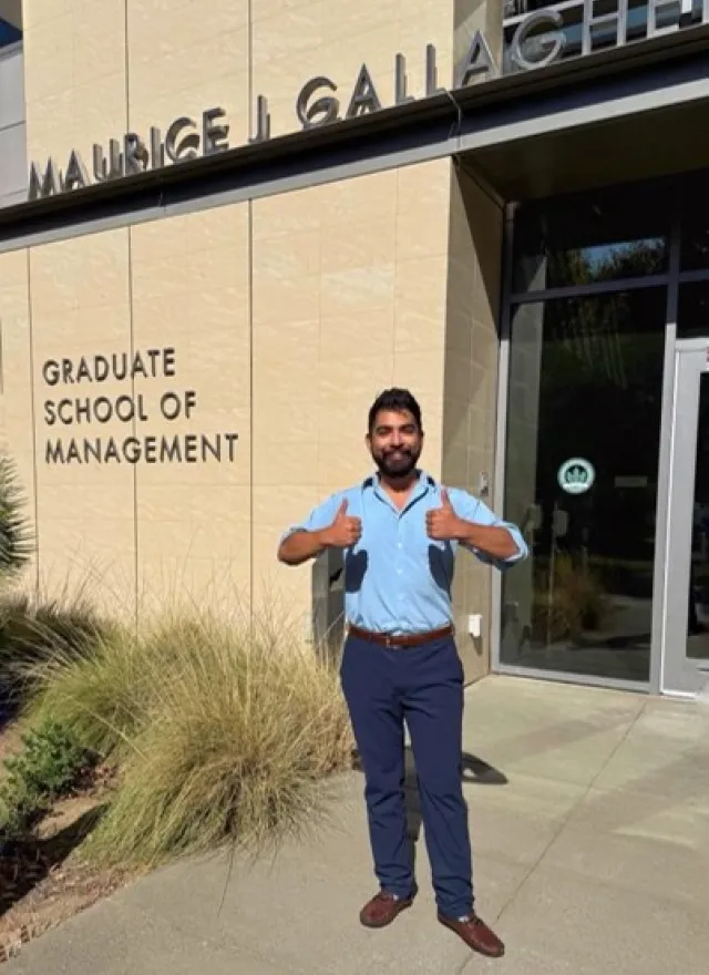 A man in a light blue shirt and dark trousers stands in front of the Maurice J. Gallagher Graduate School of Management building giving two thumbs up.