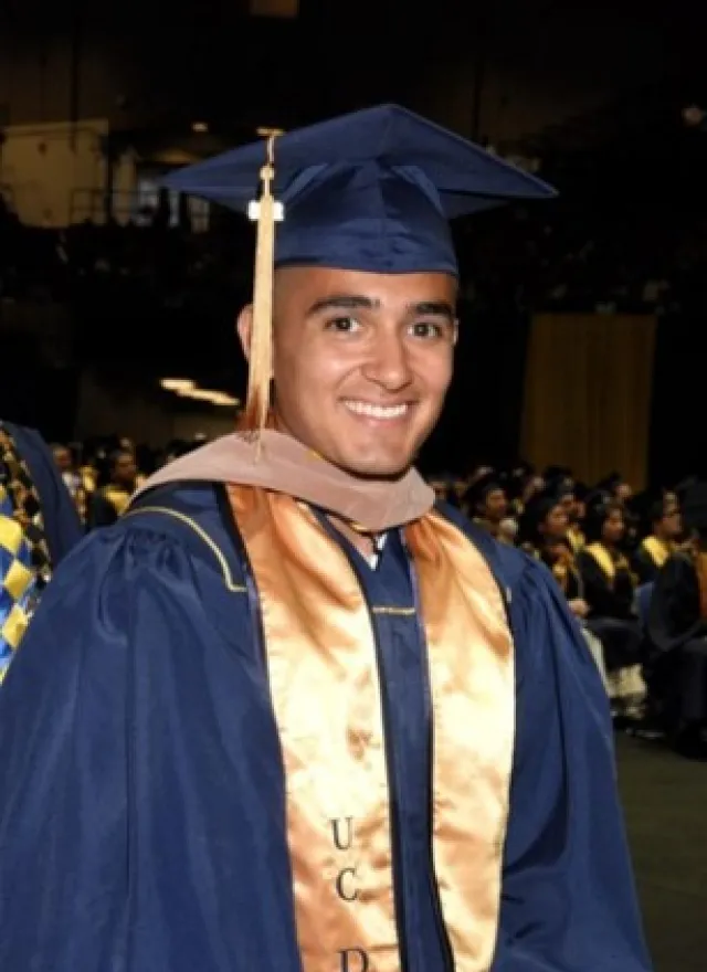 A smiling graduate in a blue cap and gown with a gold stole at a commencement ceremony.