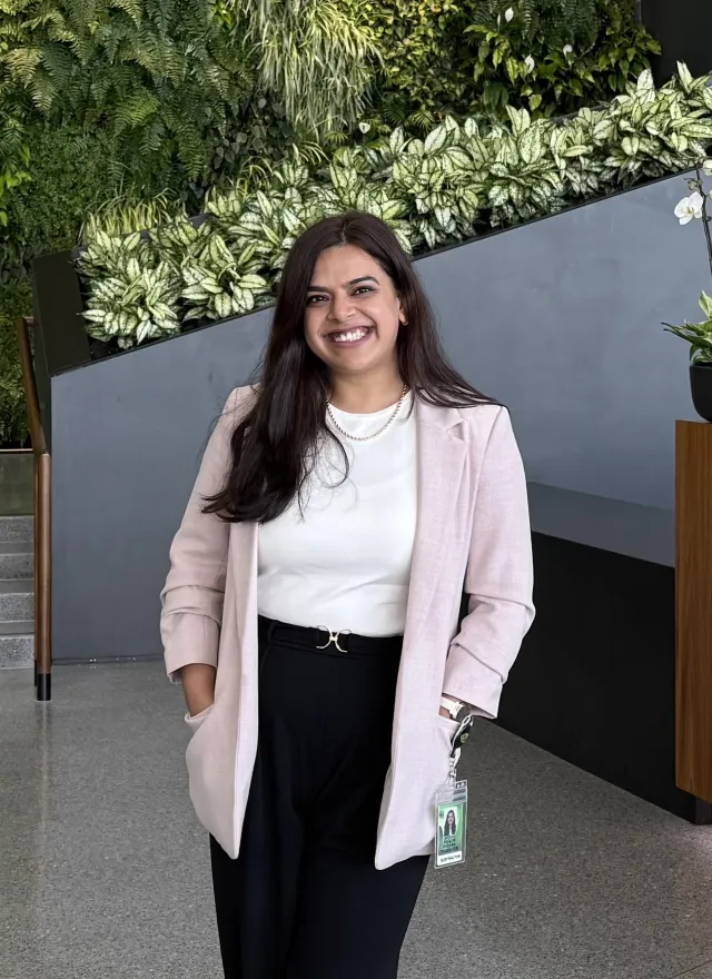 A smiling woman wearing a light pink blazer and white top stands in front of a plant-covered wall.