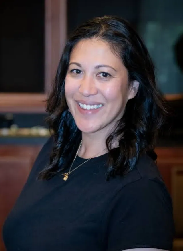 A woman with dark hair smiling at the camera, wearing a black shirt and a gold necklace.