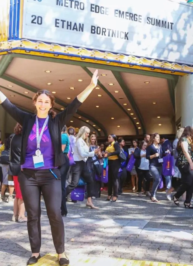 A woman with a lanyard stands smiling with arms raised in front of a theater as a crowd exits; the marquee reads "Metro Edge Emerge Summit 20 Ethan Bortnick.