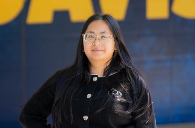 Jenny Nguyen stands in front of a blue and yellow background, wearing a black sweater with decorative buttons.