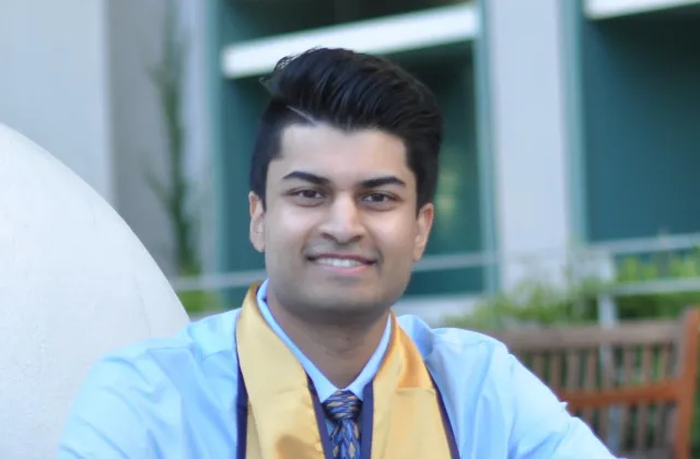 A person wearing a light blue shirt, patterned tie, and a gold graduation stole is smiling outdoors in front of a building.