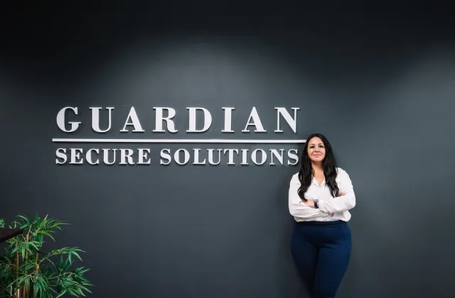 A woman stands with arms crossed in front of a dark wall displaying the words "Guardian Secure Solutions.