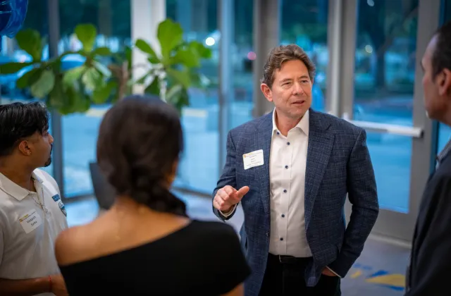 A man in a suit jacket talks to three people at an indoor event, wearing a name tag, with large windows and plants in the background.