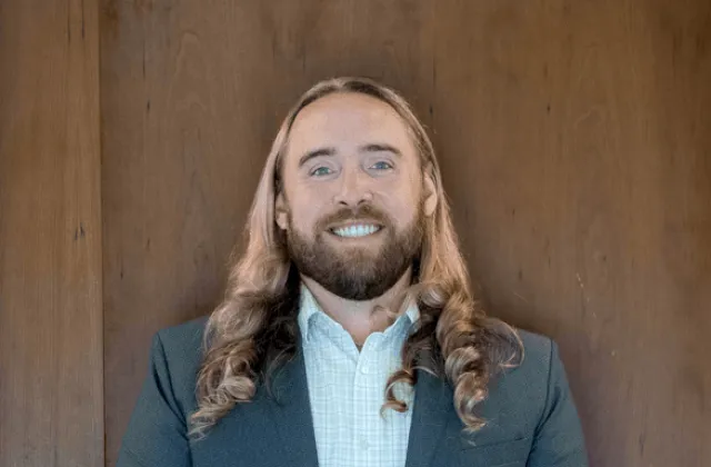 A person with long blond hair and a beard, wearing a suit jacket and checked shirt, stands smiling in front of a wooden background.