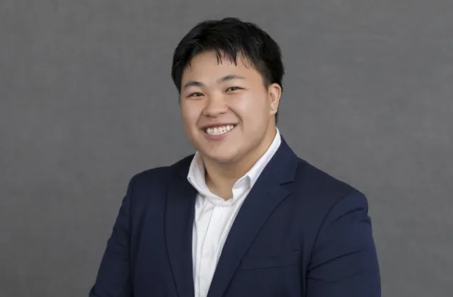 Person in a navy suit and white shirt smiling in front of a plain gray background.