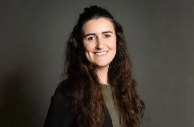 A woman with long dark hair smiles at the camera against a grey background.
