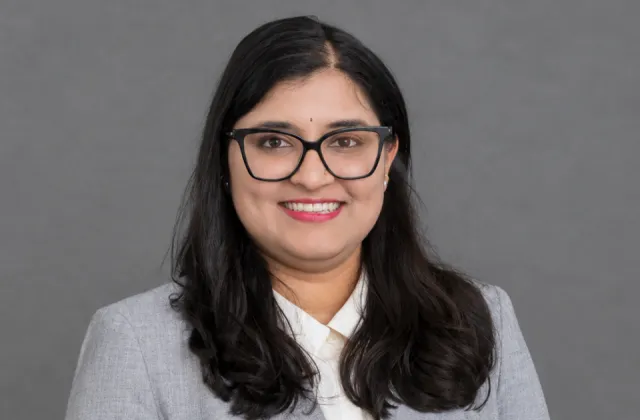 A professional headshot of a smiling woman with dark hair and glasses wearing a grey blazer over a white collared shirt.