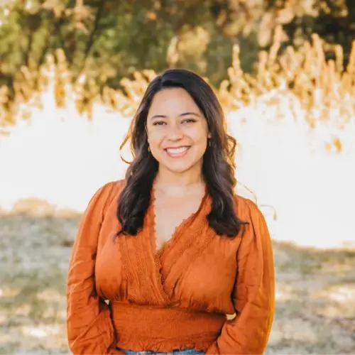 A woman with long dark hair, wearing an orange blouse, stands outdoors smiling, with blurred greenery and sunlight in the background.