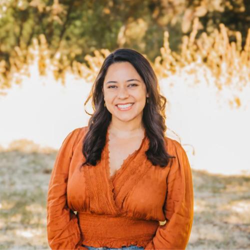 A woman with long dark hair, wearing an orange blouse, stands outdoors smiling, with blurred greenery and sunlight in the background.