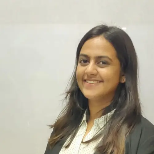 A woman with long dark hair, wearing a white blouse and black blazer, smiles while standing in front of a plain light-colored background.