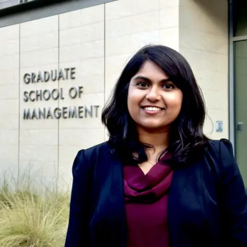 A woman in business attire stands smiling in front of a building with a sign that reads "Graduate School of Management.