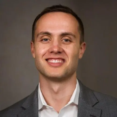 A man with short brown hair wearing a gray suit jacket and white shirt smiles at the camera against a plain background.