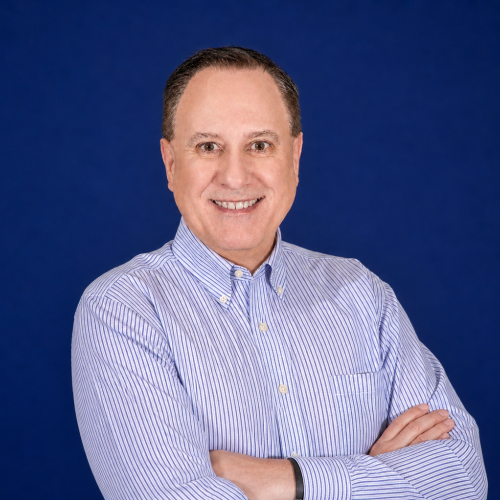 Tim Akin in a light blue striped shirt stands with arms crossed and smiles at the camera, set against a plain dark blue background.