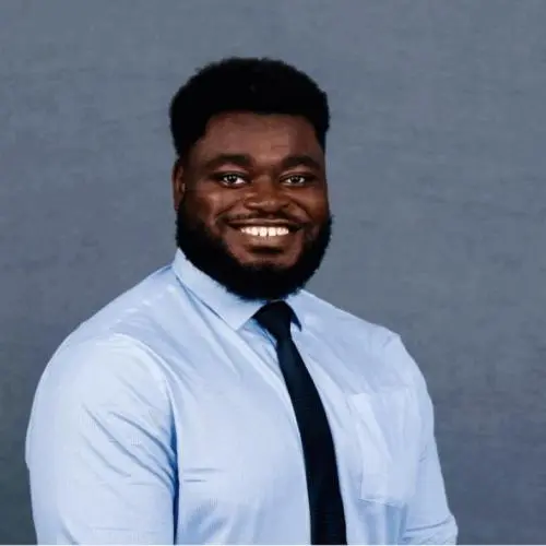 A man with a beard and afro hairstyle, wearing a light blue dress shirt and dark tie, smiles in front of a plain gray background.