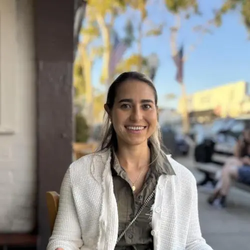 A woman with long hair, wearing a white sweater over a green shirt, sits outdoors at a restaurant table, smiling at the camera. Trees and parked cars are visible in the background.