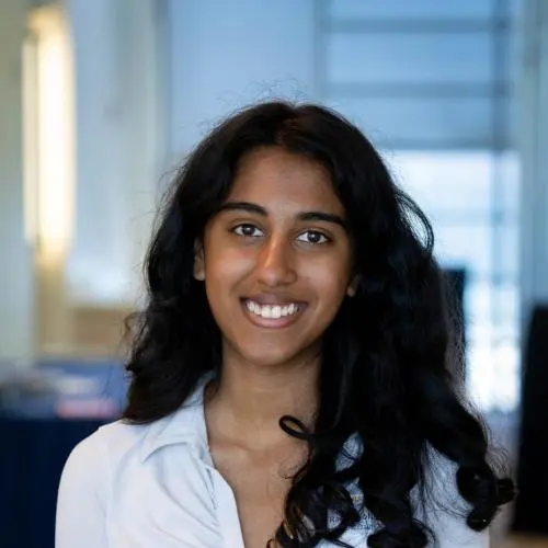 Rachel Johnson with long curly hair, wearing a white ruched blouse, stands indoors and smiles at the camera. The background is out of focus.