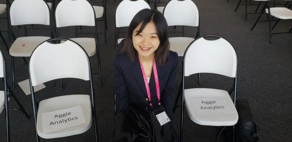 A woman in business attire sits and smiles between empty chairs labeled "Aggie Analytics" in a conference or event setting.