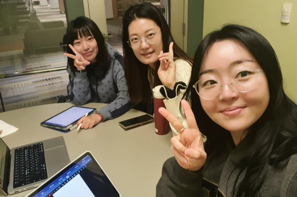 Cindy Jeon, Mia Lai, Jihyun Kim sitting at a table holding up peace signs