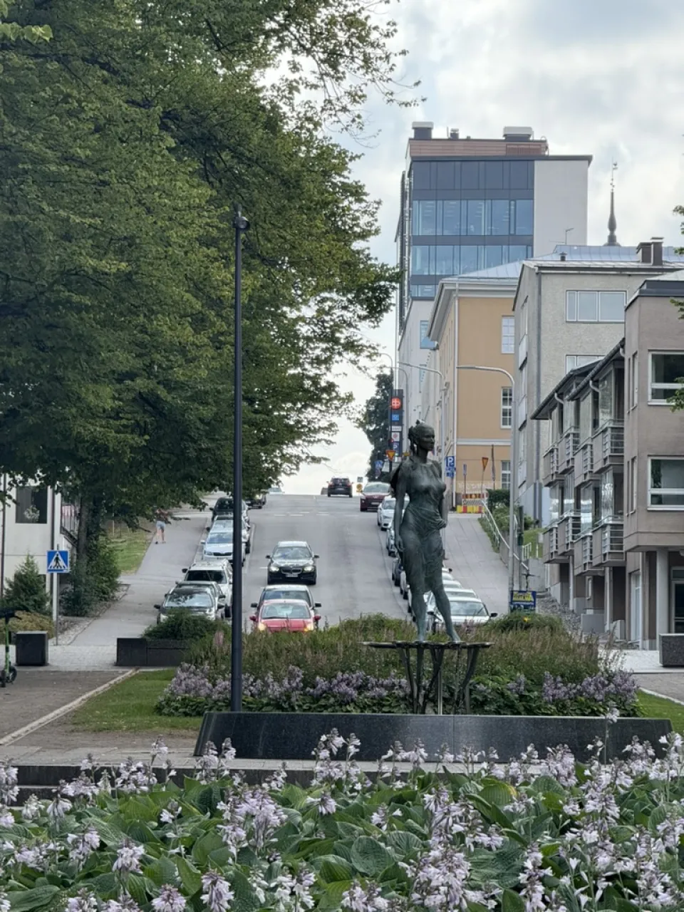 Street view in Lappeenranta, Finland, featuring a bronze statue in the foreground, flowerbeds, and a tree-lined road leading uphill past modern and historic buildings.