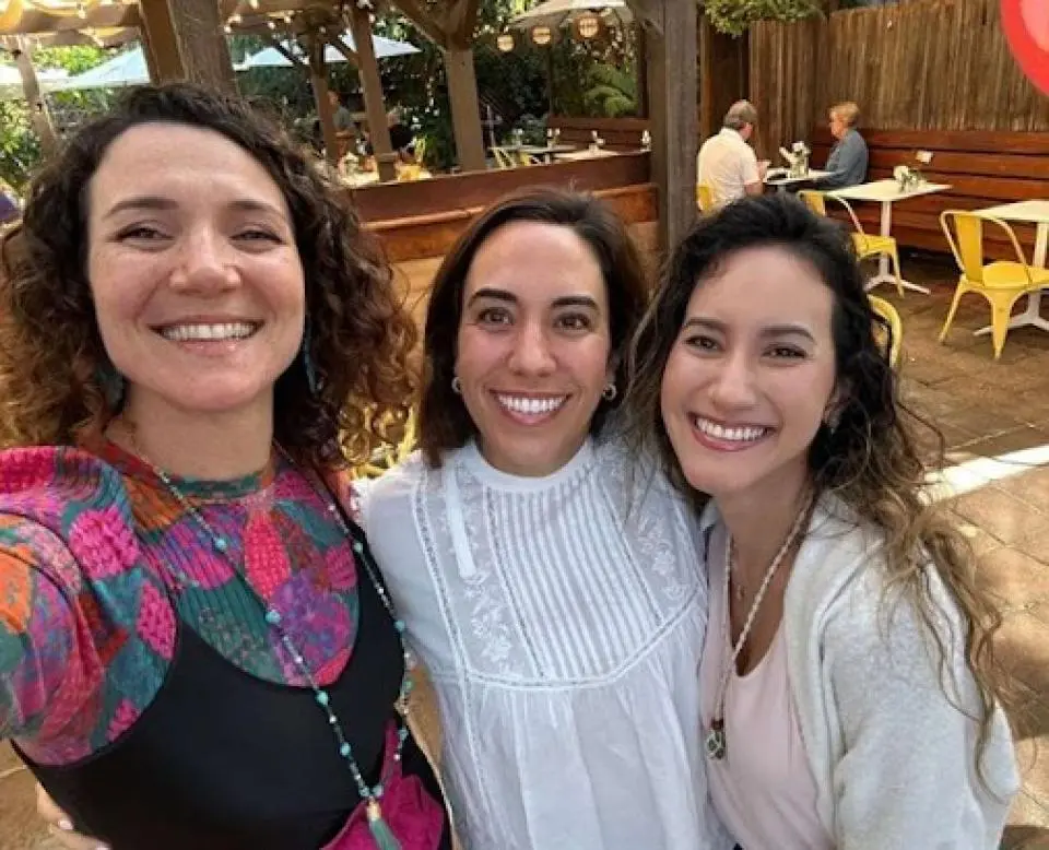 Old Novoa with two women, smiling outdoors at a restaurant patio, taking a selfie together. 