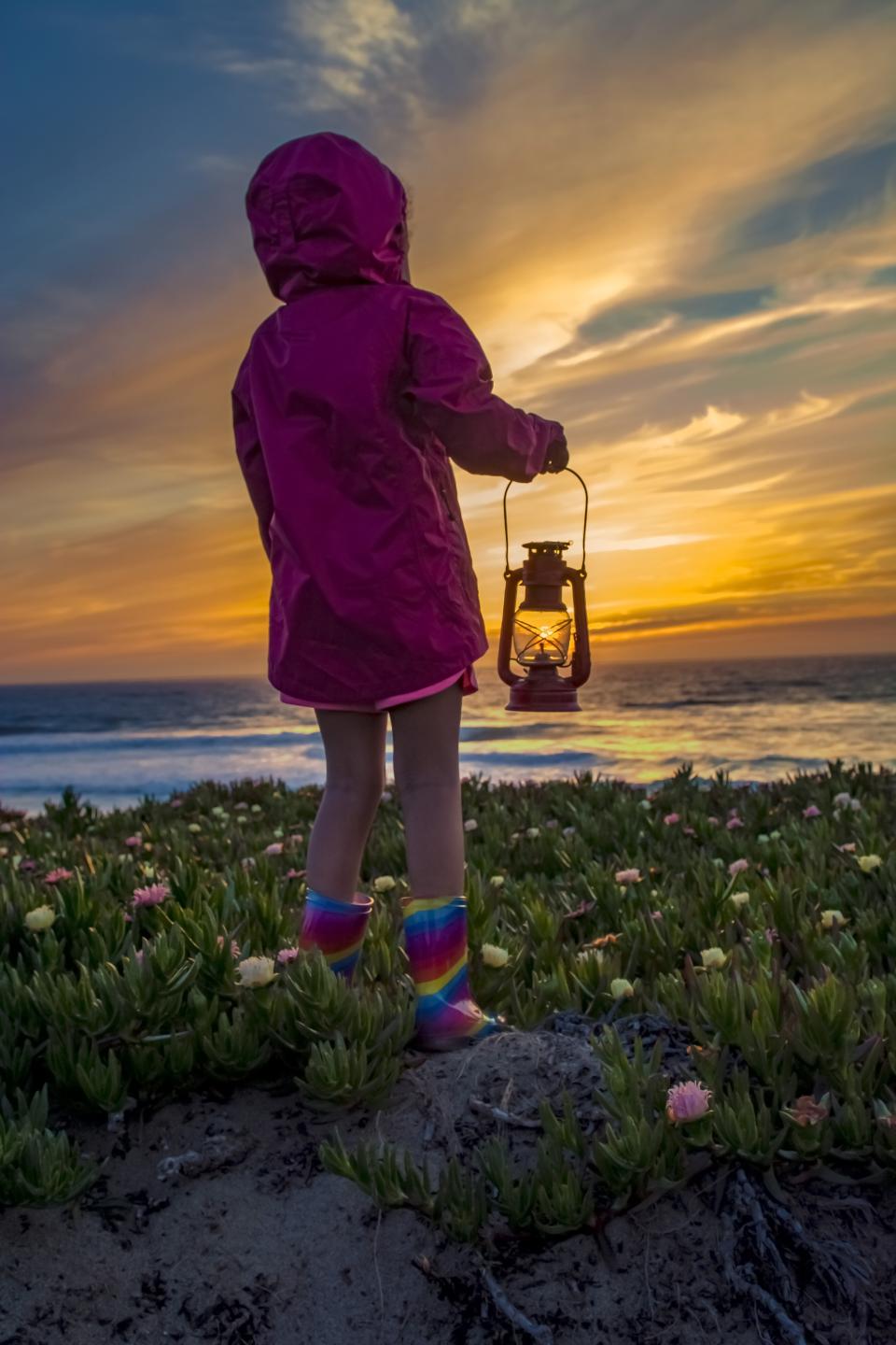 Child in a pink raincoat and rainbow boots holding a lantern while standing on a coastal bluff at sunset, facing the ocean.