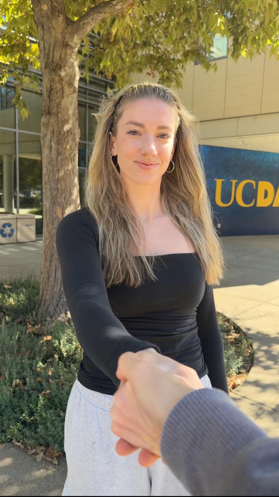 smiles and shakes hands with someone outside Gallagher Hall, standing near a tree with fall leaves.