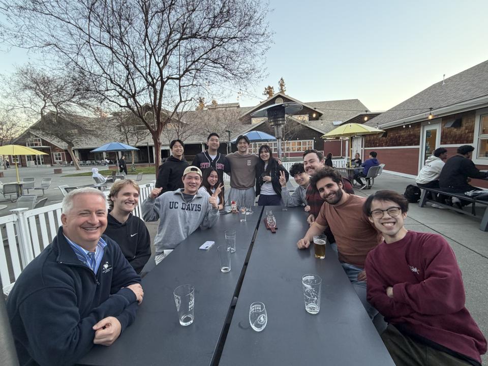 A group of people sit and stand around outdoor picnic tables at a restaurant or café, smiling at the camera; empty glasses and drinks are on the tables.