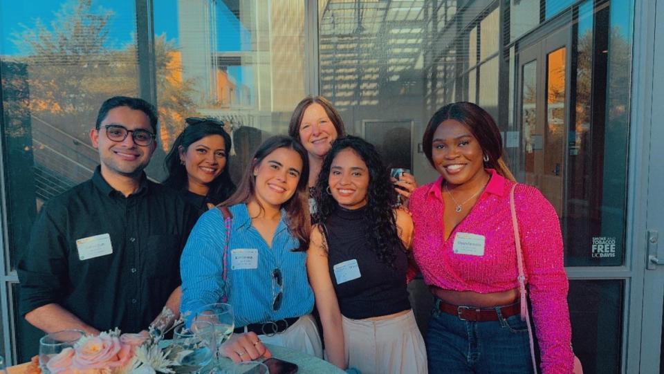 Six people stand together outdoors, smiling at the camera, each wearing name tags. There are drinks and flowers on the table in front of them. Glass windows and sunlight are in the background.
