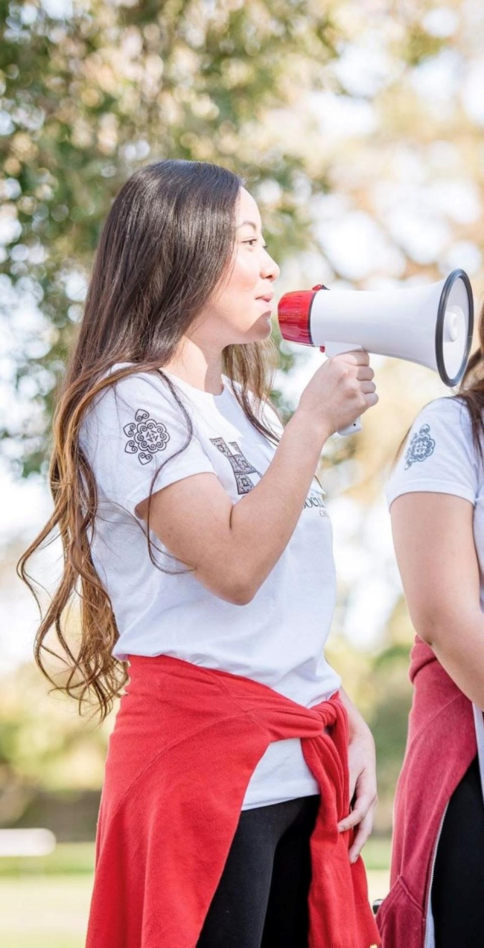 A person with long hair holds a megaphone and speaks outdoors, wearing a white t-shirt, black pants, and a red sweatshirt tied around the waist.