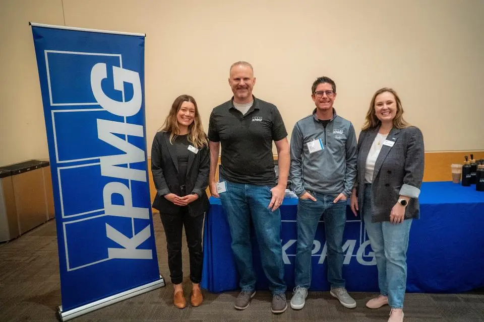 Four people stand and smile beside a KPMG banner and table in a conference room, wearing business casual attire and name tags.