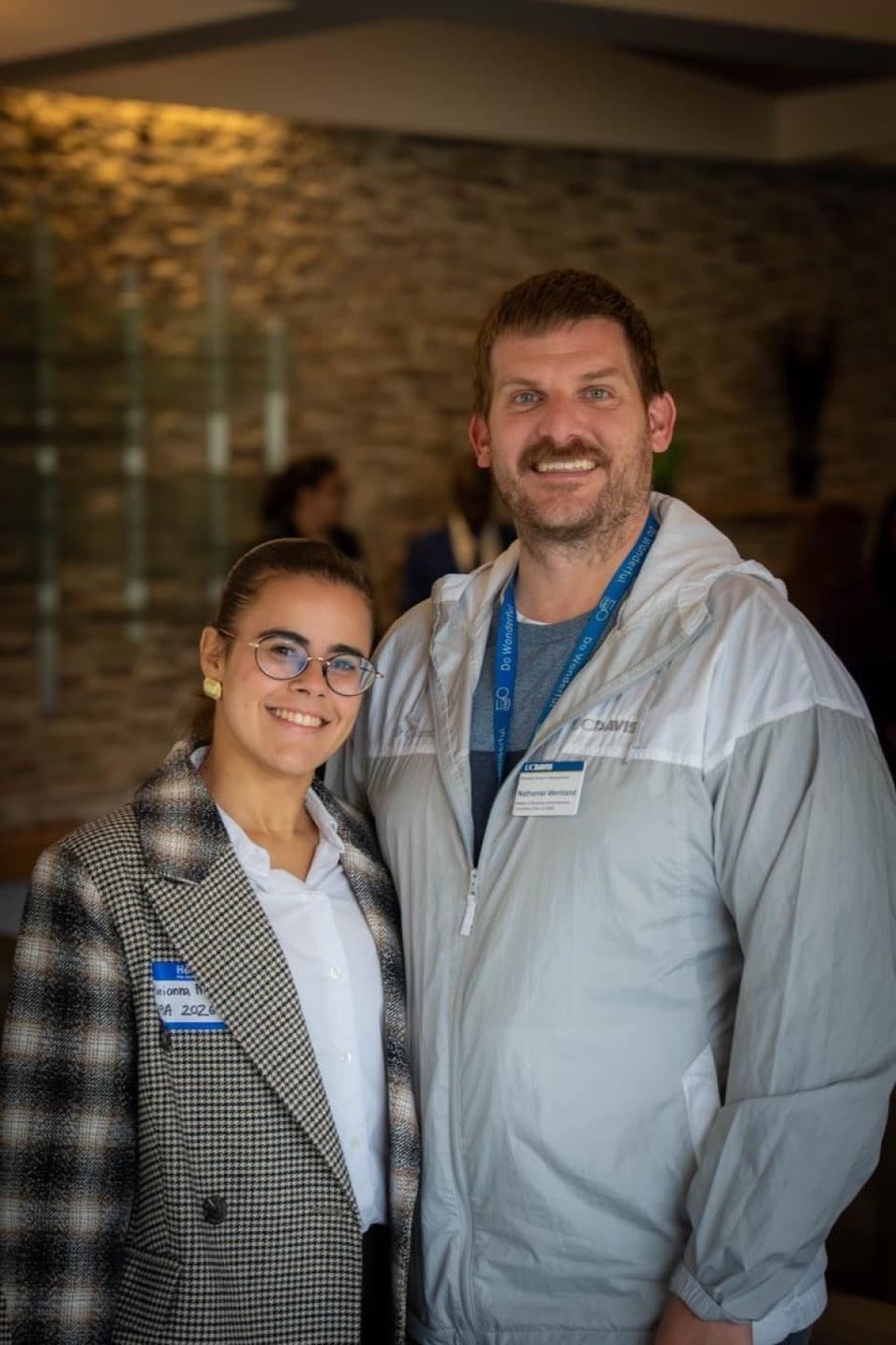 Two people standing indoors and smiling at the camera, both wearing name tags and business casual attire. The background is softly blurred with several people visible.