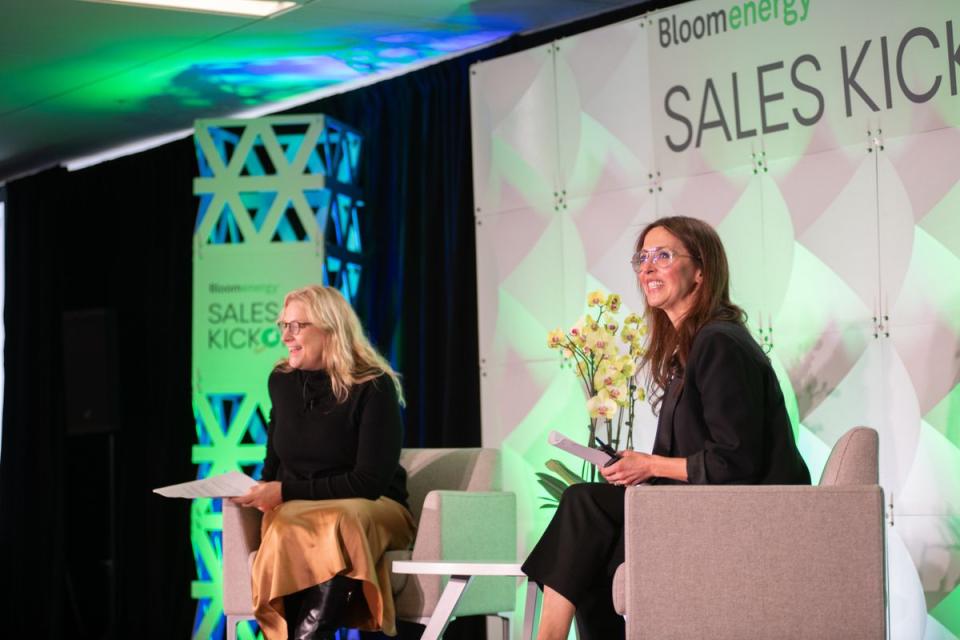 Two women sit on stage chairs with papers in hand at a Bloom Energy SALES KICKOFF event; a branded backdrop and green lighting are visible.