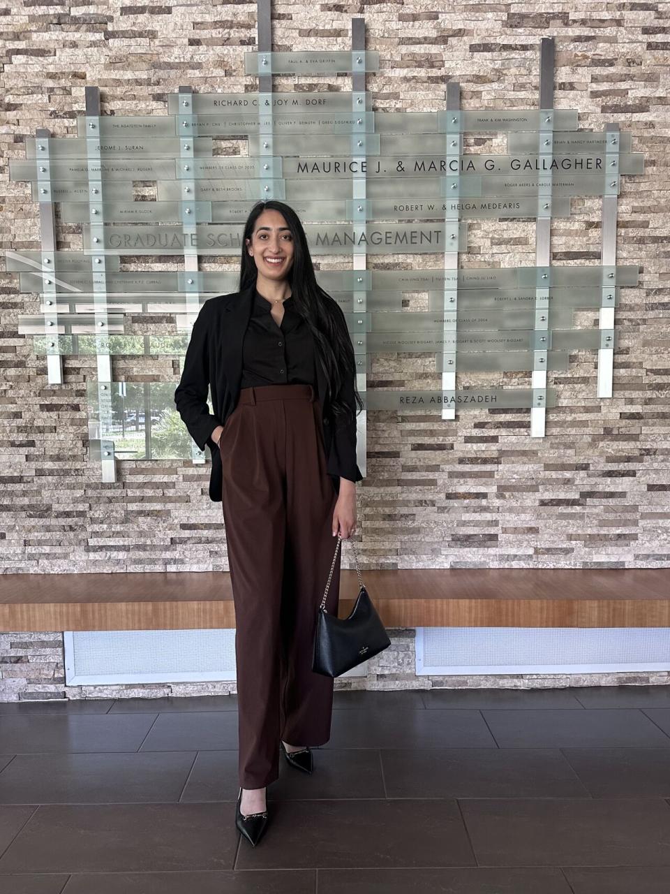 A woman stands indoors in business attire, holding a handbag and smiling, in front of a wall displaying names and a sign reading “Graduate School of Management.”