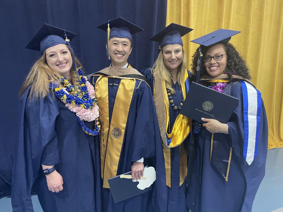 Four graduates in blue and gold caps and gowns pose together, smiling, holding diplomas and certificates, with a blue and yellow curtain in the background.
