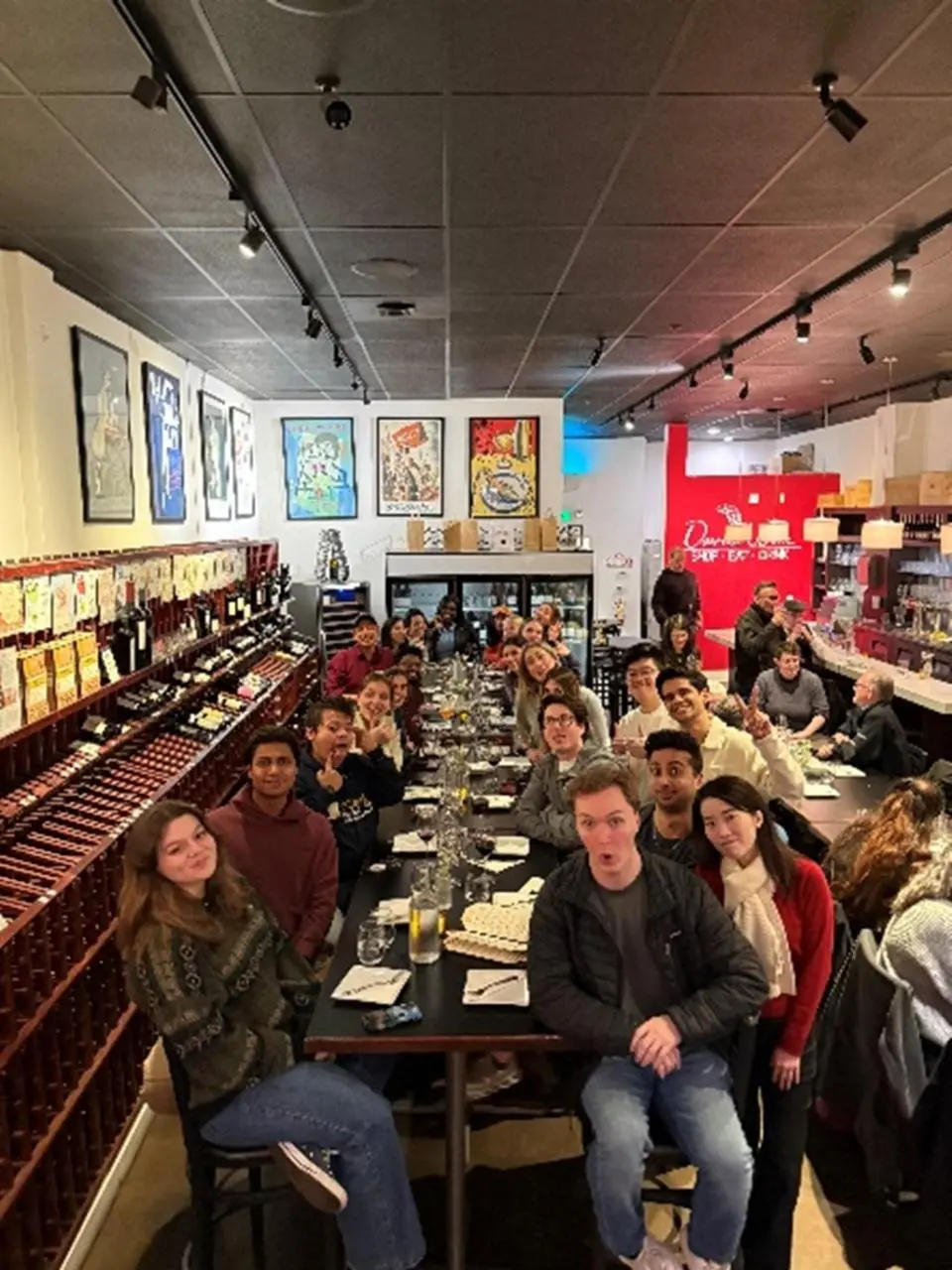 A large group of people sits around a long table in a restaurant, posing for a photo. Wine bottles line one wall, and colorful artwork hangs in the background.