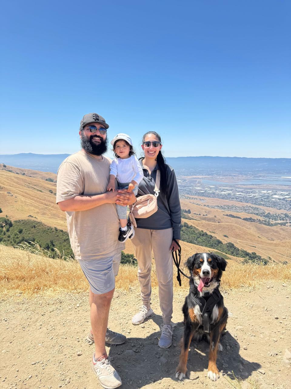 A man, woman, child, and a dog stand on a hilltop trail with a view of valleys, water, and distant mountains under a clear blue sky.