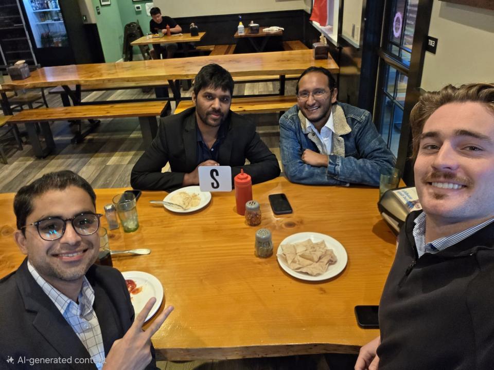 Four men sitting around a wooden table at a restaurant, smiling at the camera; snacks and drinks are on the table.
