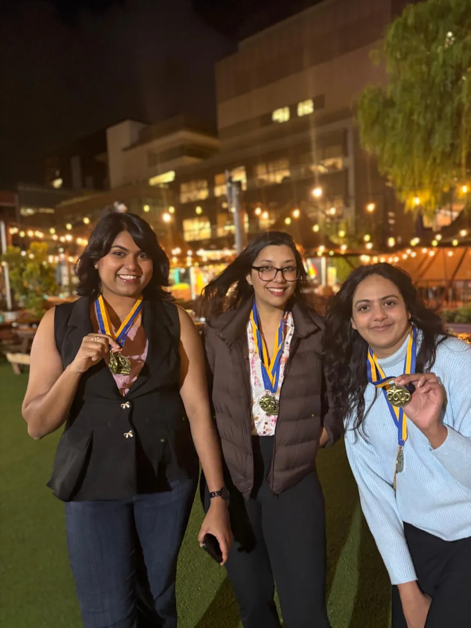 Three women stand outdoors at night, smiling and holding up medals on ribbons around their necks, with string lights and buildings in the background.