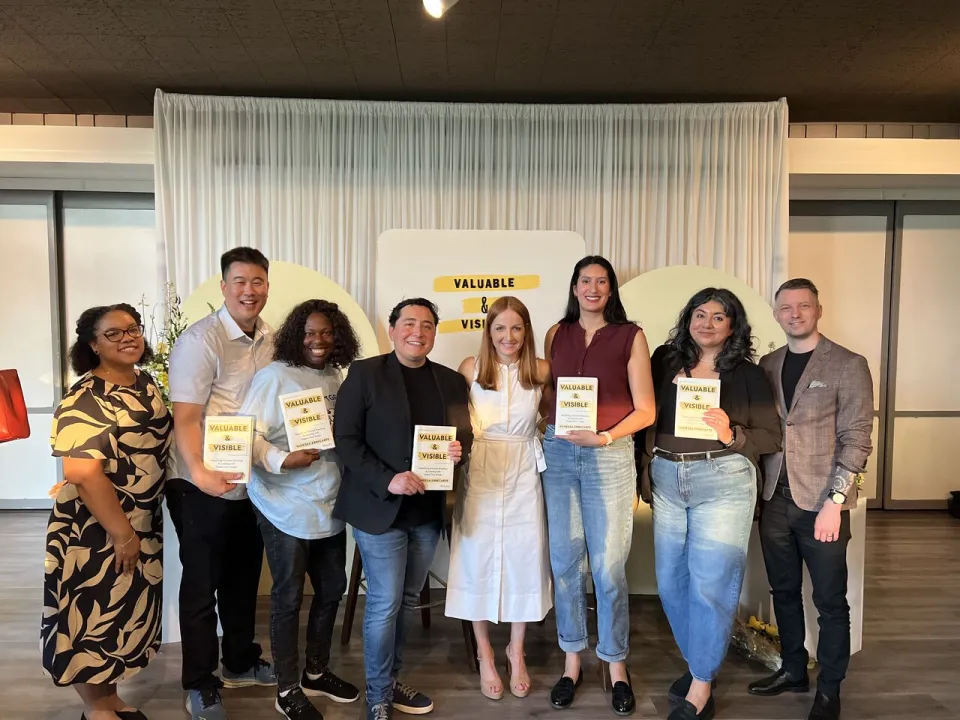 Eight people stand in a row indoors, holding copies of a book titled "Valuable & Visible," smiling at the camera with a sign and white curtain backdrop behind them.