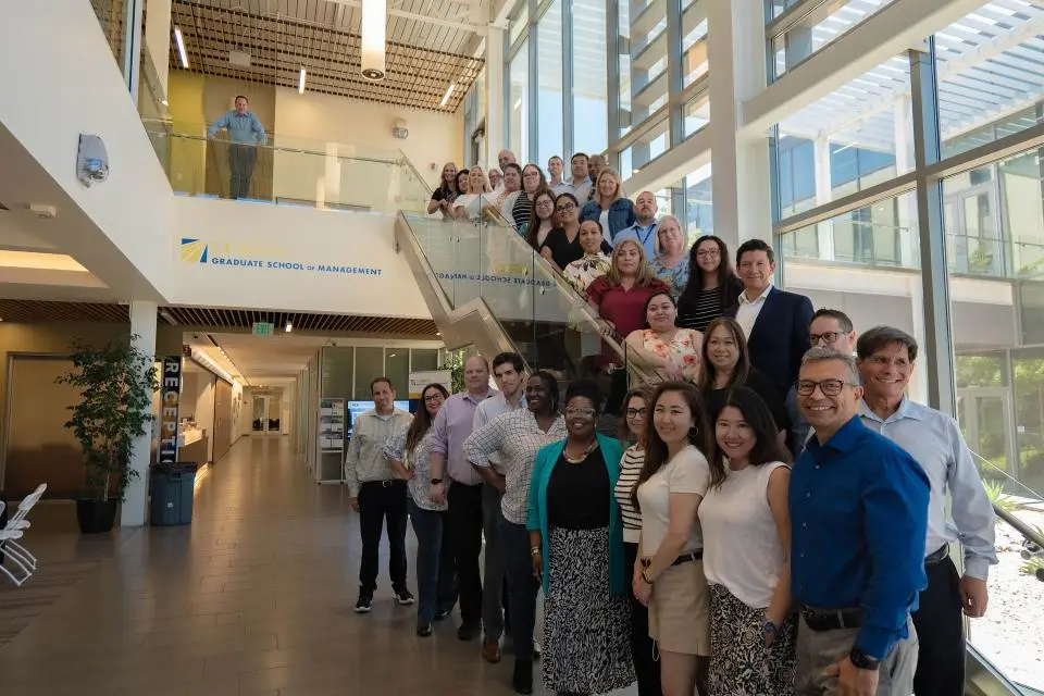 California Leadership Academy participants and speakers stand in a line on and beside a staircase inside a modern building with glass walls and natural light.