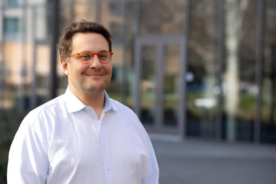 Olivier Rubel stands outside in front of a glass building, looking at the camera and smiling.