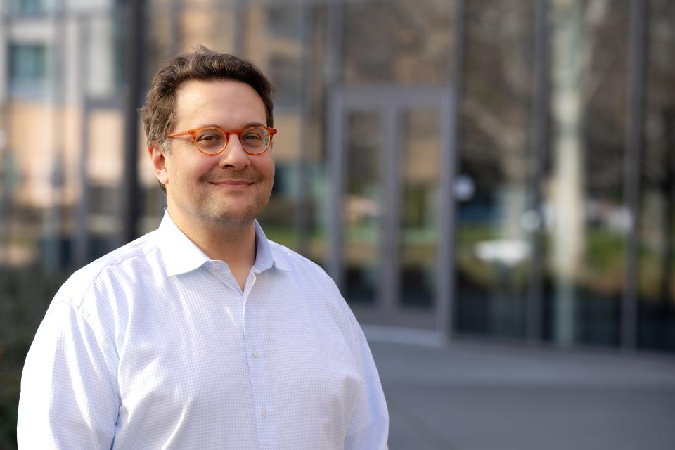 Olivier Rubel stands outside in front of a glass building, looking at the camera and smiling.