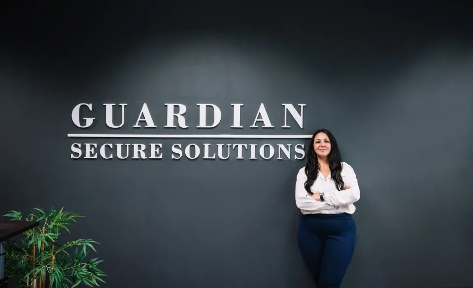 A woman stands with arms crossed in front of a dark wall displaying the words "Guardian Secure Solutions.