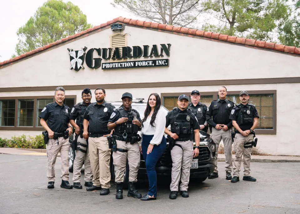 A group of security personnel in uniform stands in front of a building labeled "Guardian Protection Force, Inc." with a woman in business attire.