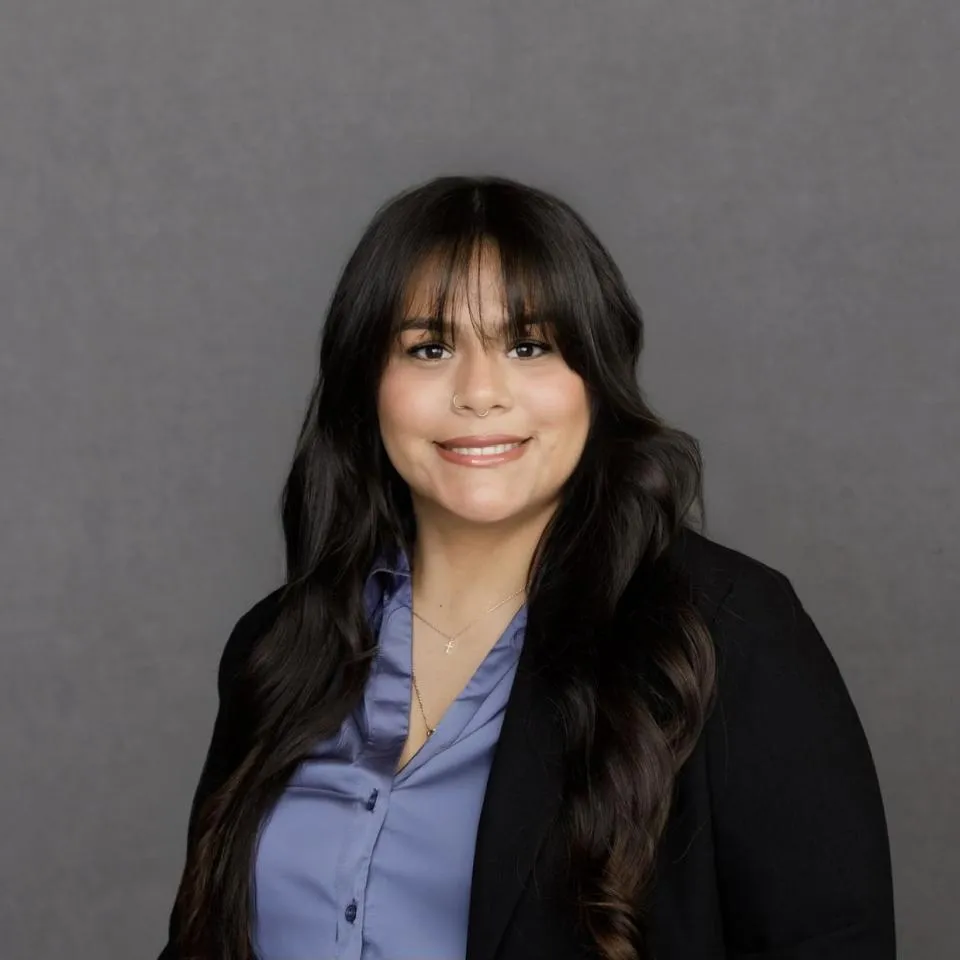 A woman with long dark hair, wearing a blue blouse and black blazer, smiles in front of a plain gray background.
